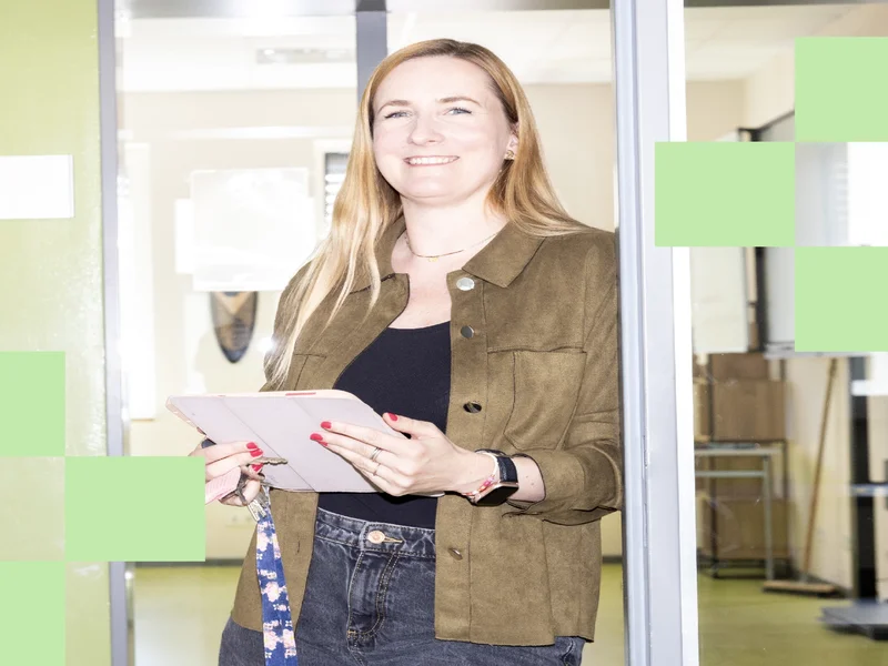 Lehrerin Ann-Kathrin Kallies lehnt an einer Tür, schaut in die Kamera und hält einen Tablet-PC in der Hand.